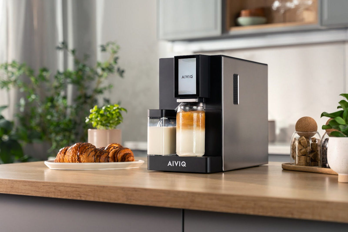 Sort espressomaskine med mælkebeholder og et glas latte på et køkkenbord ved siden af en tallerken croissanter