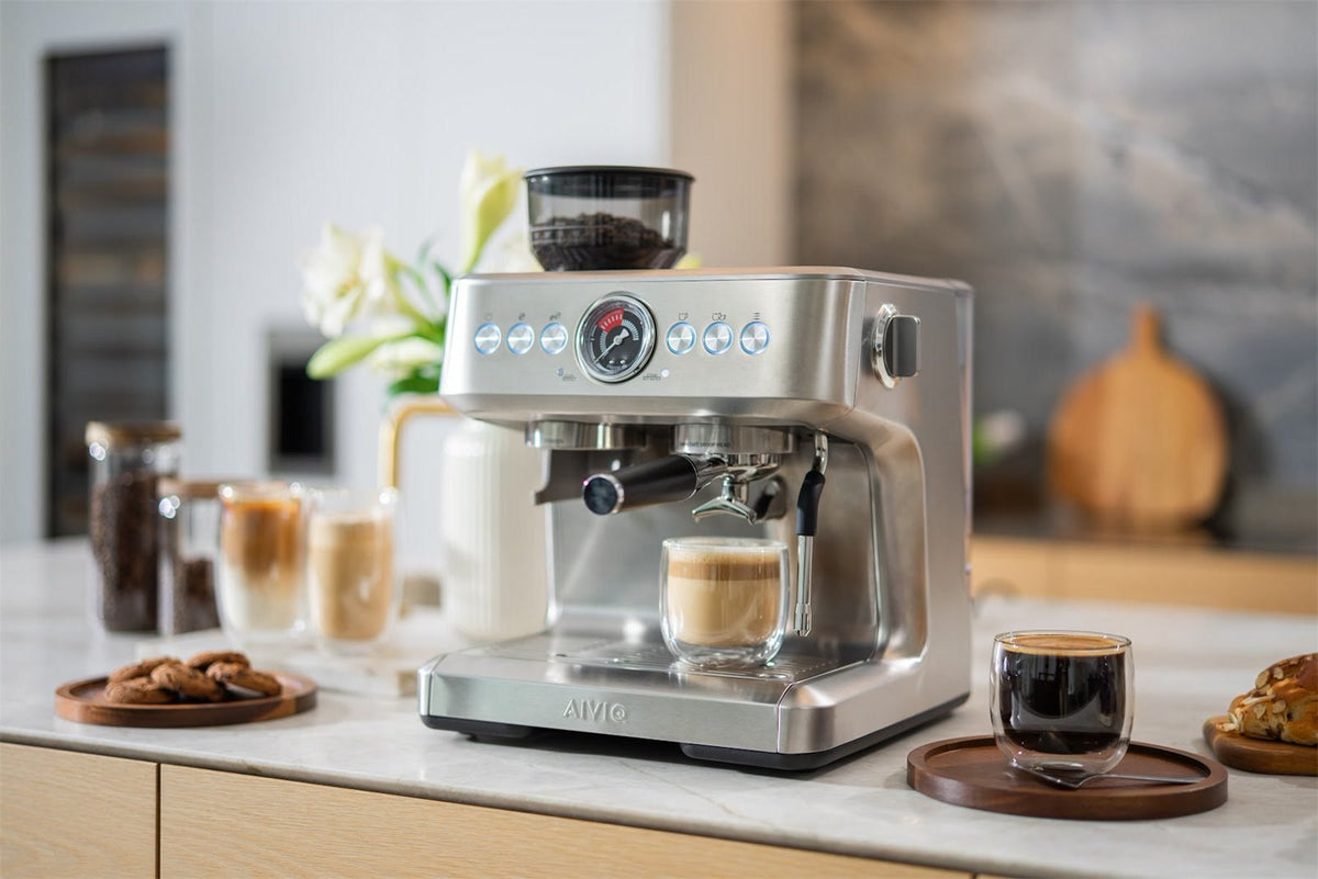 Espressomaskine i stål med bønnebeholder og manometer, et glas kaffe med mælk under tuden og en sort kaffe på et træbræt ved siden af