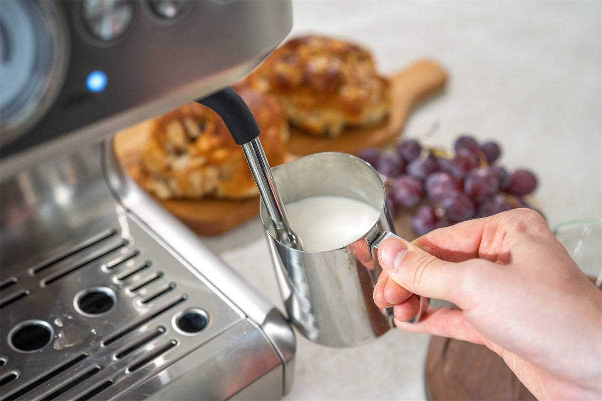 Hånd holder en stålkande med mælk under dampstaven på en espressomaskine i stål, med boller og vindruer bagved