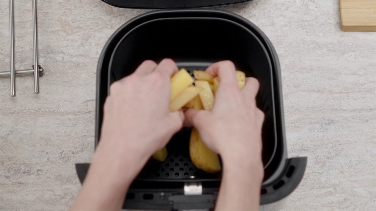To hænder lægger kartoffelbåde i kurven på en airfryer, set ovenfra