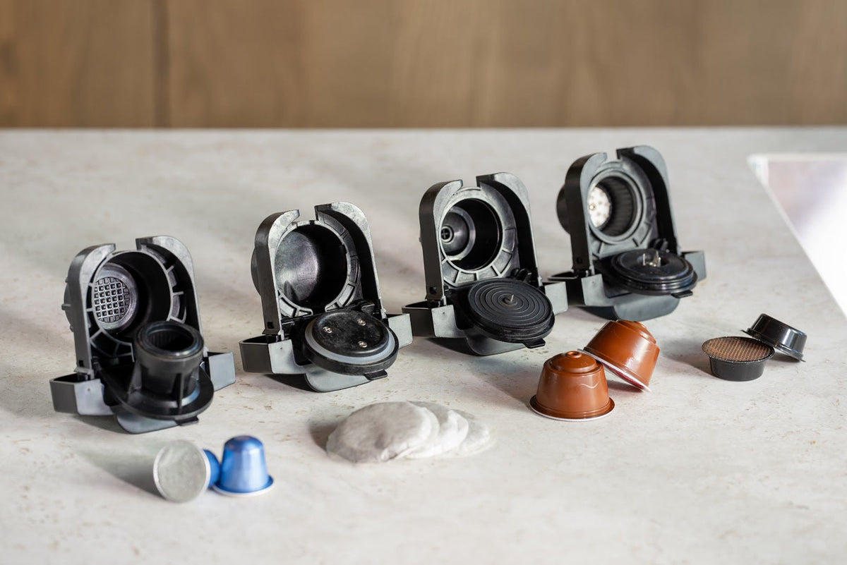 Fire åbne kapseladaptere på et bord med kapsler i forskellige former og bløde kaffepuder