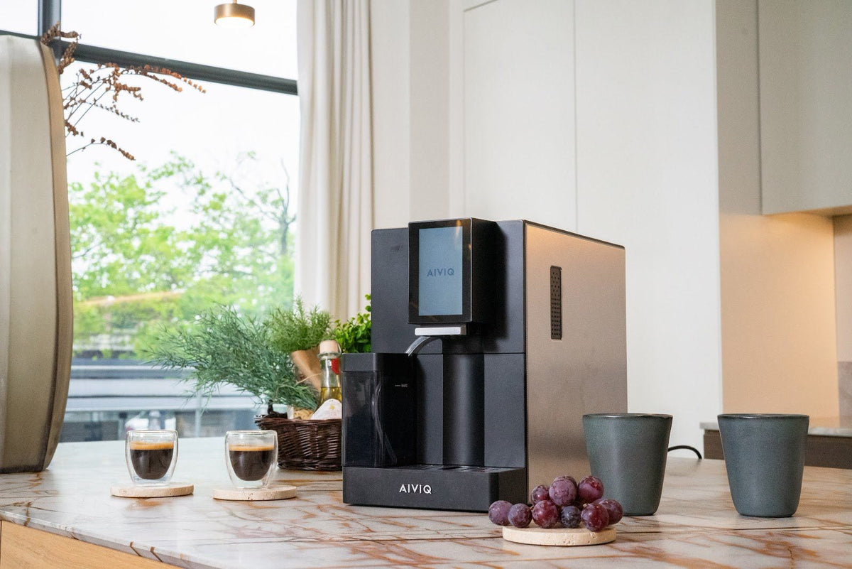 Sort espressomaskine med touchskærm og mælkebeholder på et marmorbord ved et vindue, med to espressoer i glas, to krus og vindruer