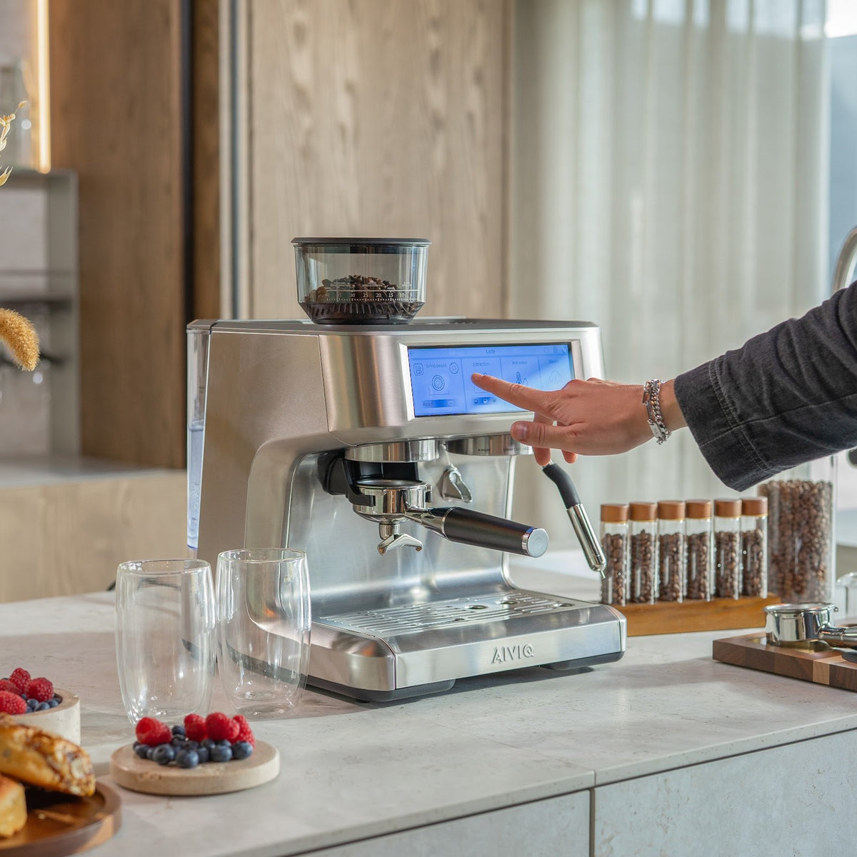 En person trykker på touchskærmen på Aspire Expert i et lyst køkken. På bordpladen står glas, bær og kaffebønner i glasrør.